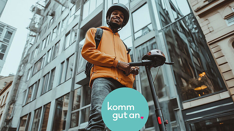 Ein junger Mann mit Helm und Rucksack steht auf einem E-Scooter in einer städtischen Umgebung mit modernen Glas- und Betongebäuden im Hintergrund. Er trägt eine orangefarbene Jacke, lächelt und blickt freundlich in die Kamera. Unten im Bild befindet sich ein rundes, türkisfarbenes Logo mit dem Text „komm gut an.“