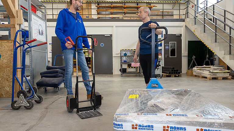 Zwei Männer stehen in der Arbeitshalle der BG Ambulanz Bremen und unterhalten sich. Der Mann rechts ist Patient, trägt schwarze Sportleidung und umfasst mit der linken Hand den Griff eines Handhubwagens. Der Mann in Jeans links, ein Ergotherapeut, hält mit der linken Hand eine eine Stapelkarre fest. 