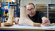 Ein Mann im schwarzen T-Shirt sitzt in einer Werkstatt und betrachtet zwei Holz-Leisten. Es sind die Muster, nach denen der Orthopädie-Schuhmachermeister die Maßschuhe fertigt. 