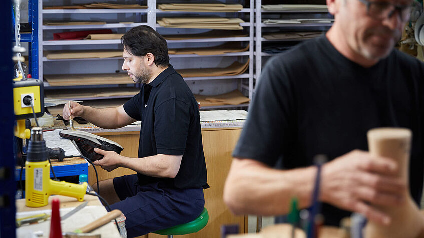 Zwei Männer arbeiten in der Werkstatt. Der Orthopädie-Schuhmacher links m Bild sitzt an einer wWerkbank und passt konfektionierte Schuhe an. Der andere Mann, rechts im Vordergrund, hat einen Holzleisten in der Hand. 