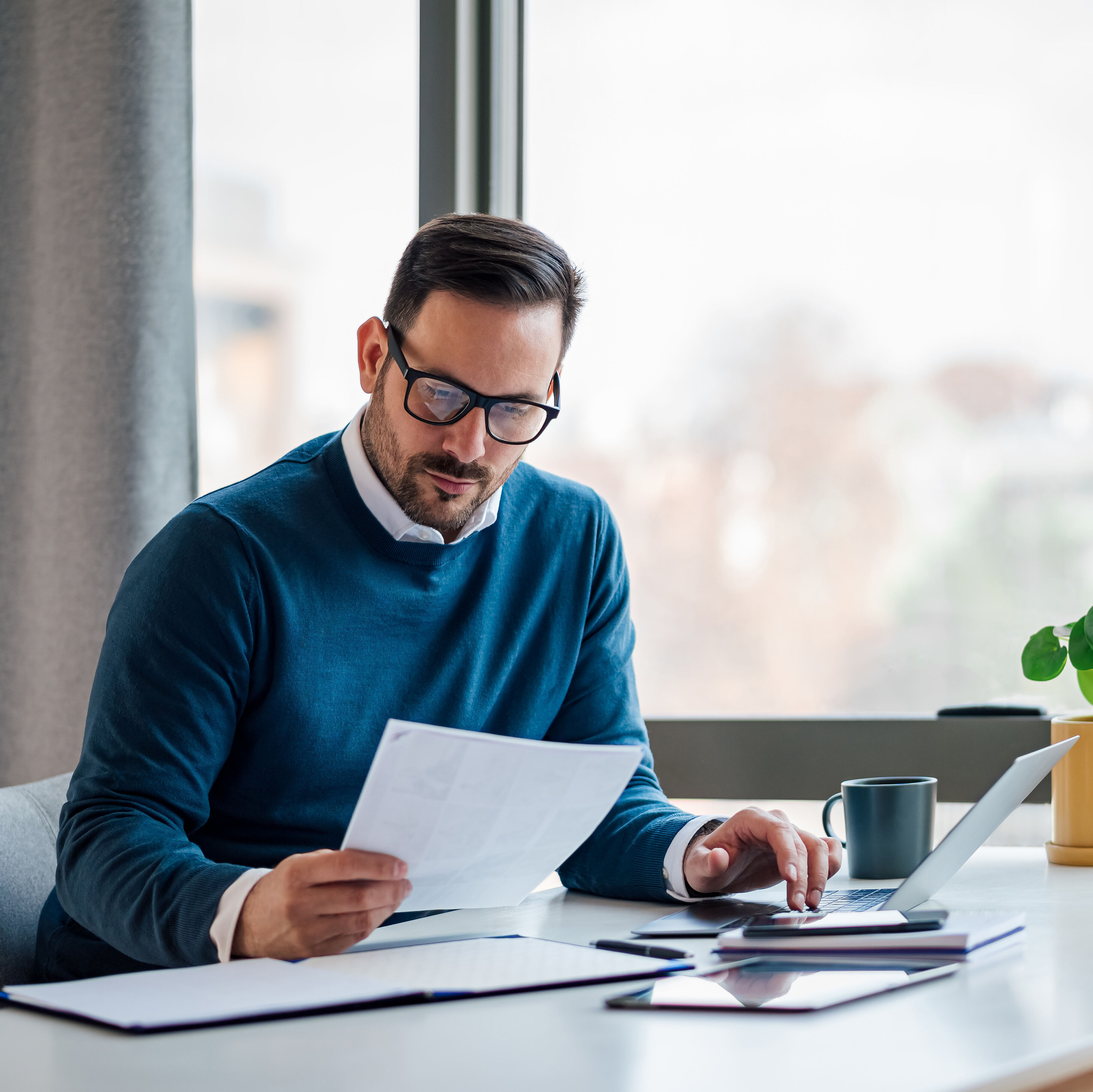 Mann liest ein Dokument und tippt auf einem Laptop, an einem Schreibtisch mit Büromaterial.