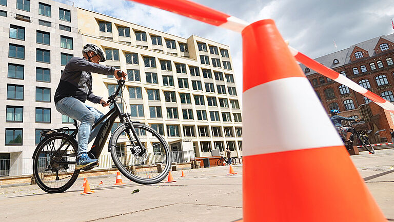 Ein Platz mit einer leuchtend rot-weiße Pylone im Vordergrund, darüber weht ein rot-weißes Flatterband. Ein Zweiradfahrer umkurvt Pylonen. Im Hintergrund sind mehrstöckige Häuserfassaden zu sehen. 