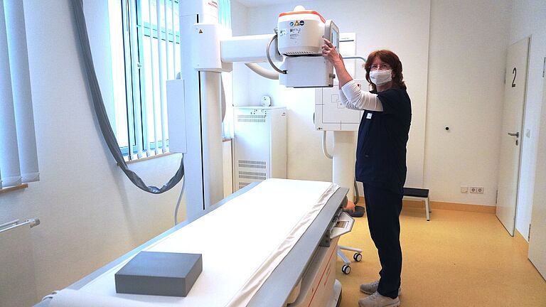 Eine Frau steht vor der Liege eines Röntgengerätes und fasst mit beiden Händen zum Röntgenkopf. Sie trägt eine Maske vor dem Mund und schaut in die Kamera. 