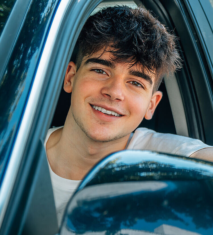 Ein junger Mann sitzt im Auto und schaut aus dem geöffneten Fenster der Fahrertür. Er lacht und sieht positiv aus.