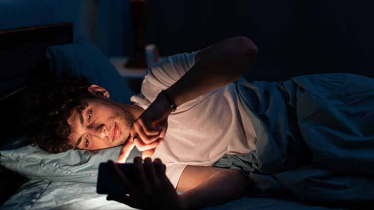 Junger Mann liegt bei Dunkelheit im Bett und schaut auf das Handy