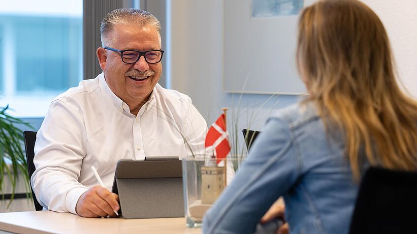 Ein Mann spricht mit einer Frau. Sie sitzen an einem Tisch. Vor dem Mann steht ein Tablet.