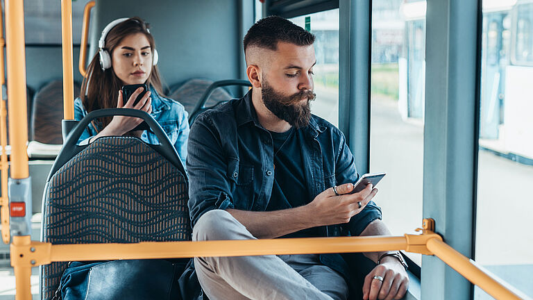 Junge Frau und junger Mann sitzen im fahrenden Bus und schauen auf ihr Handy.