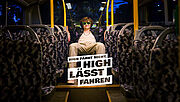 Ein junger Mann mit lockigen Haaren, Sonnenbrille und lässiger Kleidung sitzt entspannt auf der Rückbank eines leeren Busses. Auf dem Bild steht der Slogan: „High fährt nicht. High lässt fahren.“