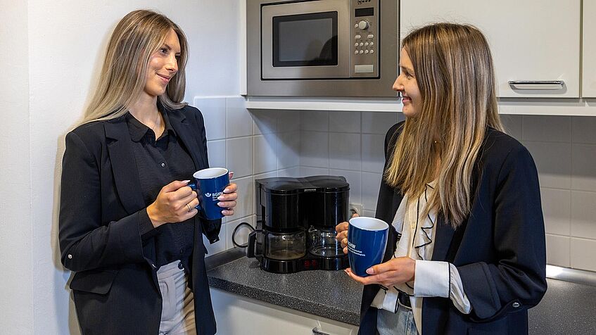 Zwei Frauen stehen in der Teeküche. Sie unterhalten sich und halten einen Teebecher in ihren Händen.
