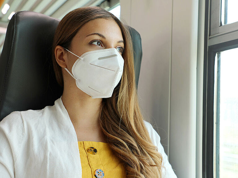 Eine Frau sitzt am Fenster eines Zuges und trägt eine FFP2-Maske.
