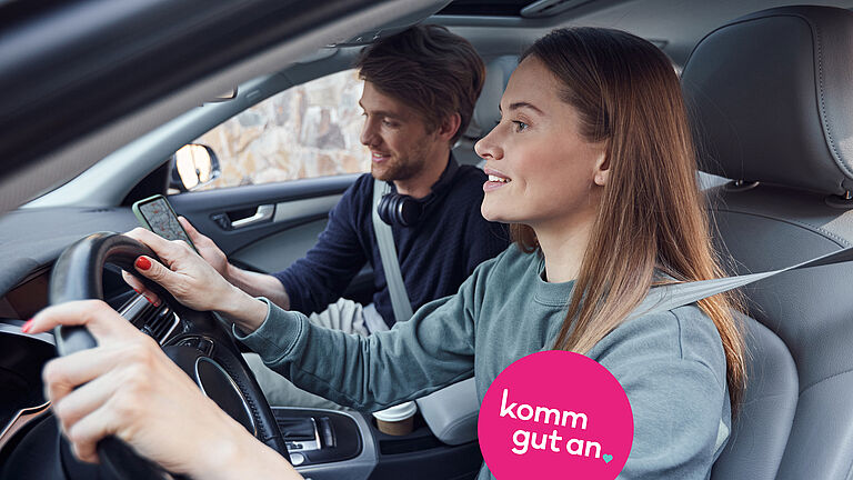 Zwei angeschnallte Personen sitzen in einem Auto. Die Person am Steuer blickt nach vorn, die Person auf dem Beifahrersitz hält ein Smartphone. Auf dem Bild ist der Schriftzug „komm gut an“ eingeblendet.