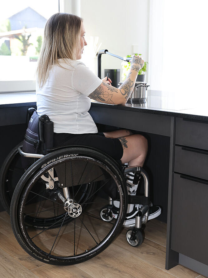 Tamara Fischer in der barrierefreien Küche ihres Hauses. Mit dem Rollstuhl steht sie am Waschbecken und füllt Leitungswasser in einen Kochtopf.