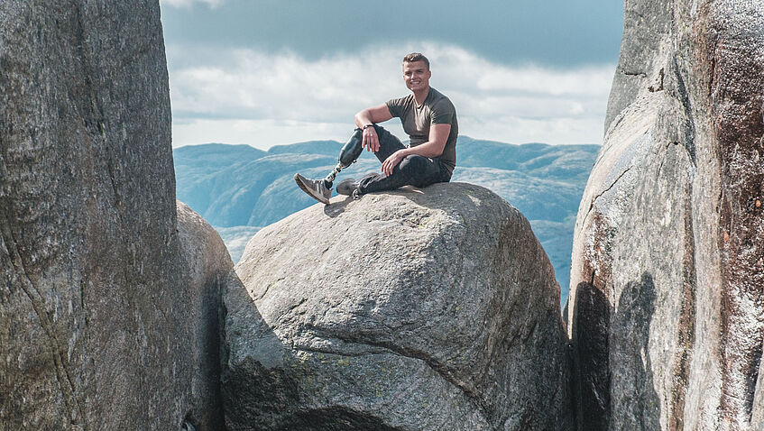 Thomas Müller, ein BGHW-Versicherter aus Köln, dessen rechter Unterschenkel nach einem Unfall amputiert werden musste, sitzt auf einem großen felsigen Stein, der zwischen zwei Felsen on der Luft hängt. n hängt und schaut in die Kamera. Er schaut freundlich in die Kamera.