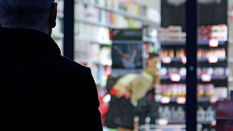 Auf dem Bild ist die Silhouette eines Mannes zu erkennen, der im Dunkeln draußen vor einem beleuchteten Geschäft steht und durch die Fensterscheibe schaut.