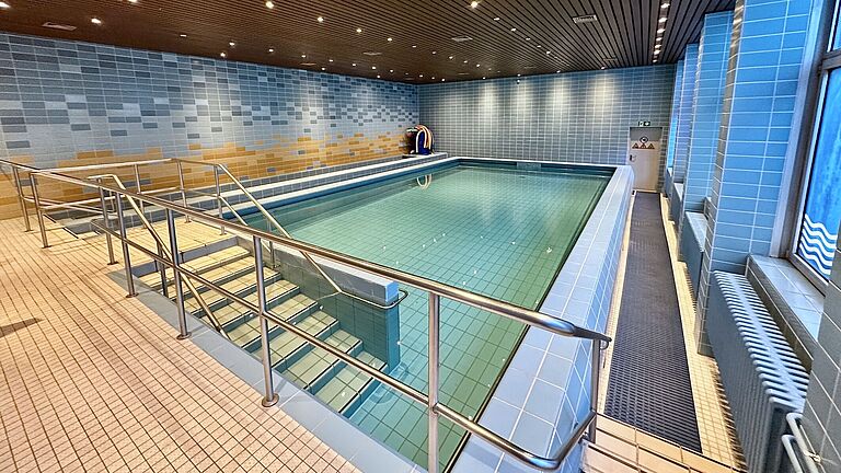 Das Foto zeigt eine große Halle mit einem Schwimmbecken in der Mitte. Eine mehrstufige Treppe führt in das Becken, die Wande sind blau gekachelt.