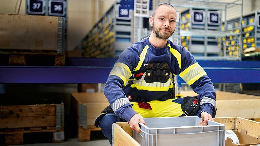 Das Bild zeigt Mitarbeiter Pascal Rudel. Er trägt ein Exoskelett und hebt eine schwere Kiste.