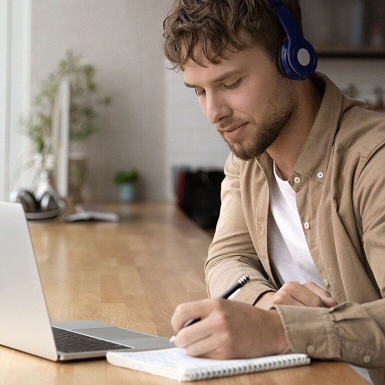 Junger Mann mit Kopfhörern sitzt vor Laptop und macht Notizen