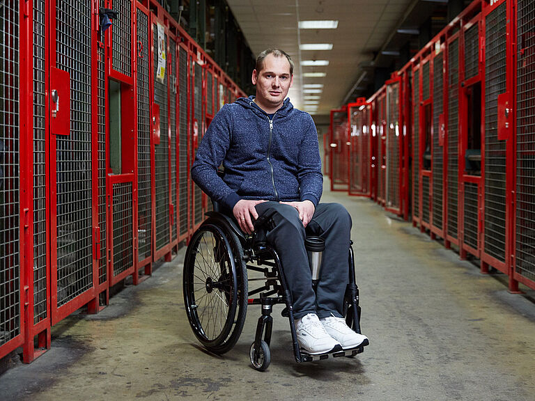 Johann Koch steht mit seinem Rollstuhl in einer Lagerhalle und lacht in die Kamera. 