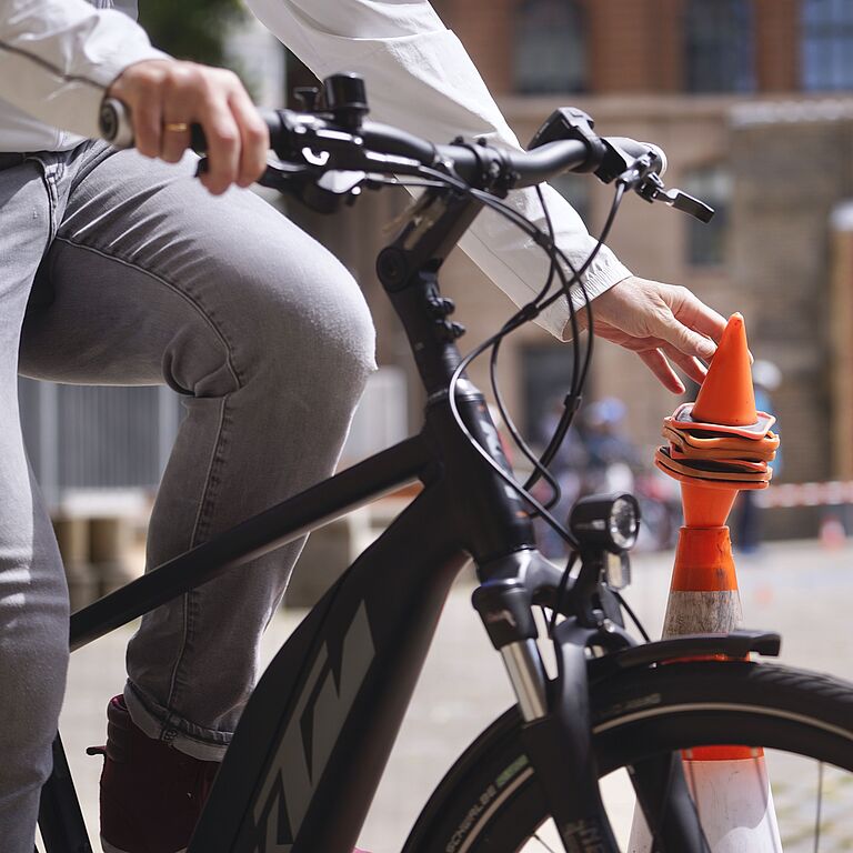 Das Bild zeigt den Ausschnitt eines schwarzen Pedelecs. Der Fahrer hält mit der linken Hand den Lenker, mit der rechten Hand setzt er ein rotes Plastikhütchen auf eine rot-weiße Pylone. 