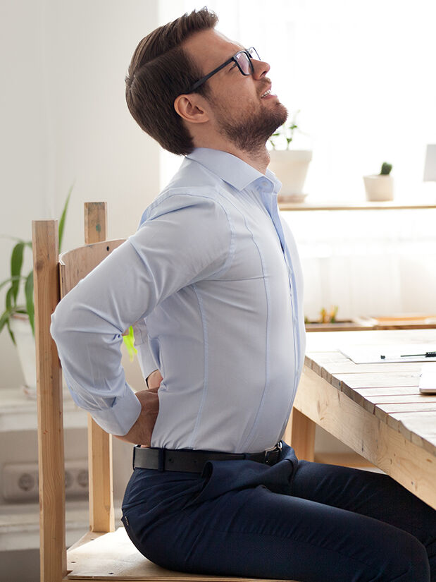 Ein Mann sitzt auf einem Holzstuhl vor einem Holztisch. Er greift sich mit den Händen an seinen schmerzenden Rücken.