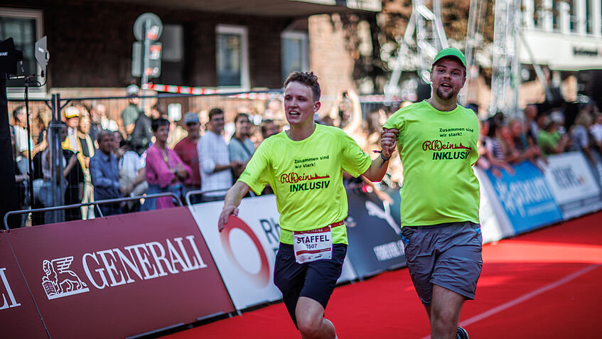 Zwei Menschen laufen in einem Staffellauf Hand in Hand. Einer von ihnen ist beeinträchtigt. Der Läufer rechts i Bild hat einen fehlgebildeten, kurzen Arm. Die Hand hält sein Laufpartner. Ober der andere Arm fehlt oder hinter dem Rücken ist, sieht man auf dem Bild nicht. Am Rand der abgesperrten Straße stehen viele Zuschauer. 