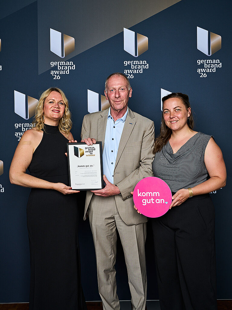 Drei Personen stehen vor einer Fotowand des German Brand Award 2026 und lächeln in die Kamera. Die Person links und der Mann in der Mitte halten gemeinsam eine Urkunde mit der Auszeichnung „German Brand Award Winner 2026“ für die Kampagne „komm gut an“. Die Person rechts hält ein rundes, pinkfarbenes Schild mit der Aufschrift „komm gut an.“. Alle drei sind festlich gekleidet; im Hintergrund ist mehrfach das Logo des German Brand Award zu sehen.