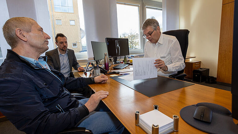 Drei Männer sitzen um einen Schreibtisch. Der Mann in weißem Hemd rechts liest in Papieren, die er in seinen Händen hält und spricht zu den gegenüber sitzenden Personen.. 