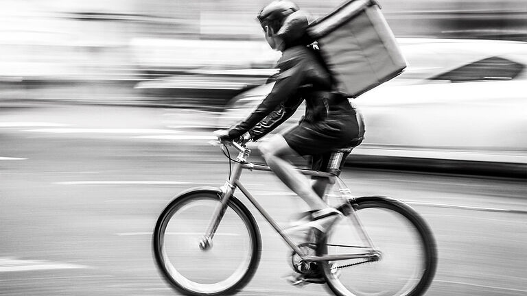 Ein Radfahrer fährt mit einem roten Mountain-Bike und einem roten Lieferrucksack über eine Straße. Im Hintergrund sieht man verwischte Autos. Der Radfahrer trägt Helm, kurze Hosen und eine schwarze Jacke. 