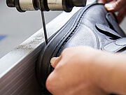 Ein Schuh, der von zwei Händen gehalten wird, liegt auf einem Metallband. Über ihm hängt ein schmales Bohrmesser, das die Schuhsohle längs auftrennt. 